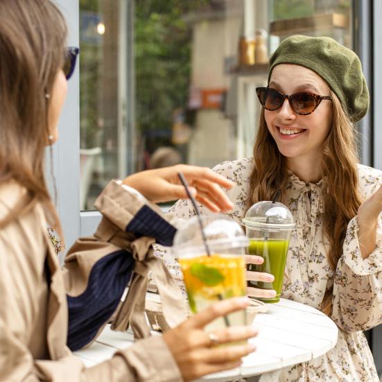 To kvinder, der taler sammen. De sidder udenfor på en café og drikker juice eller smoothie.