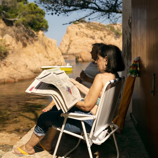 Kvinde, der læser avis i en havestol udenfor.