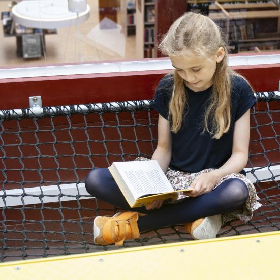En pige, der sidder i et edderkoppenet og læser en bog i børnebiblioteket.