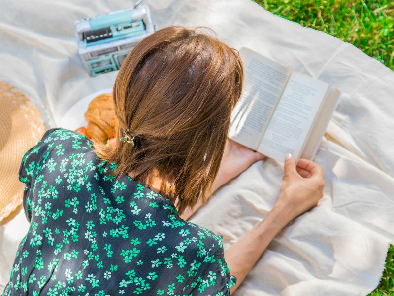 Kvinde, der lægger på et tæppe og læser en bog udenfor.